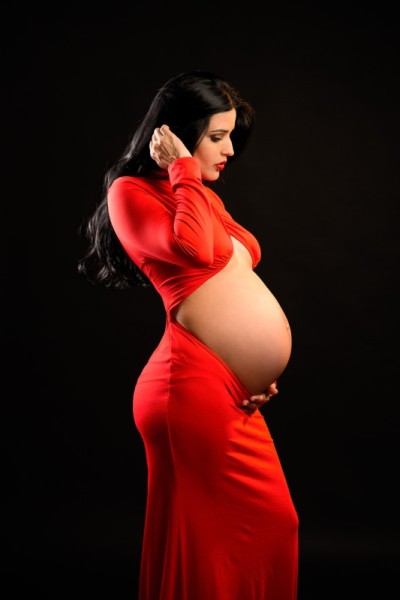 Studio maternity photo of pregnant woman in elegant red dress holding her belly in profile, with dramatic lighting on dark background